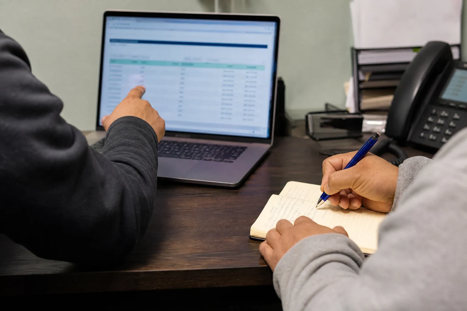 Over-the-shoulder view of one person pointing at a table on a laptop screen while another writes notes in a notebook at a wooden office desk with a desk phone.