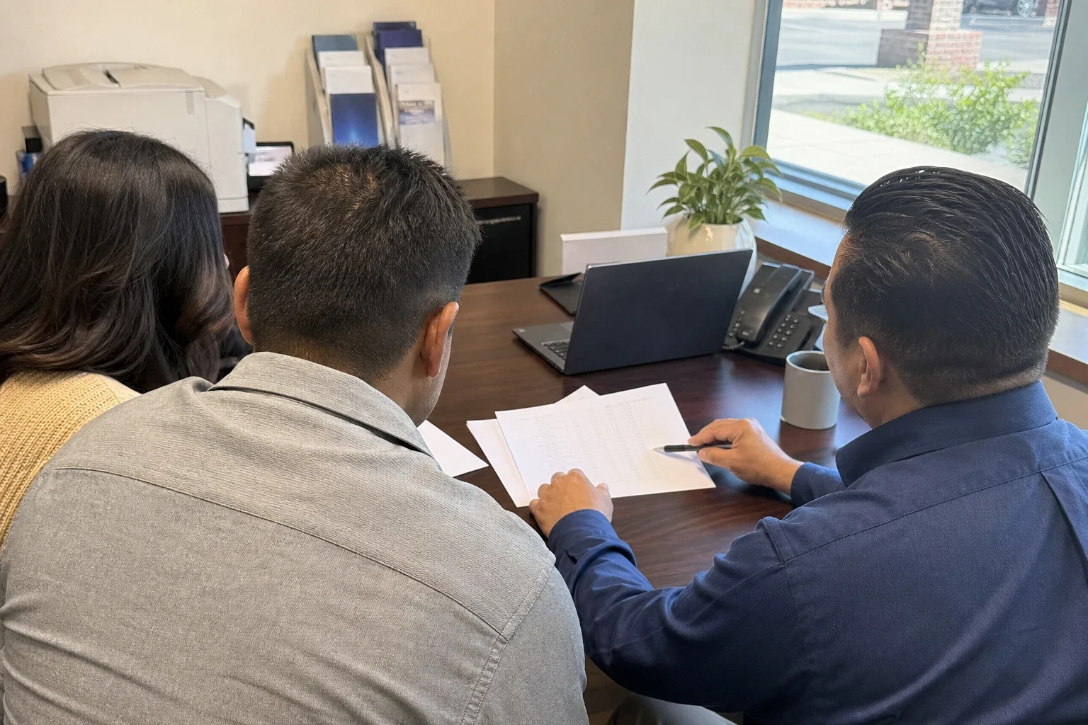 Over-the-shoulder view of a couple at an office desk as an agent in a blue shirt points a pen at a printed table of numbers, with a laptop, desk phone and window nearby