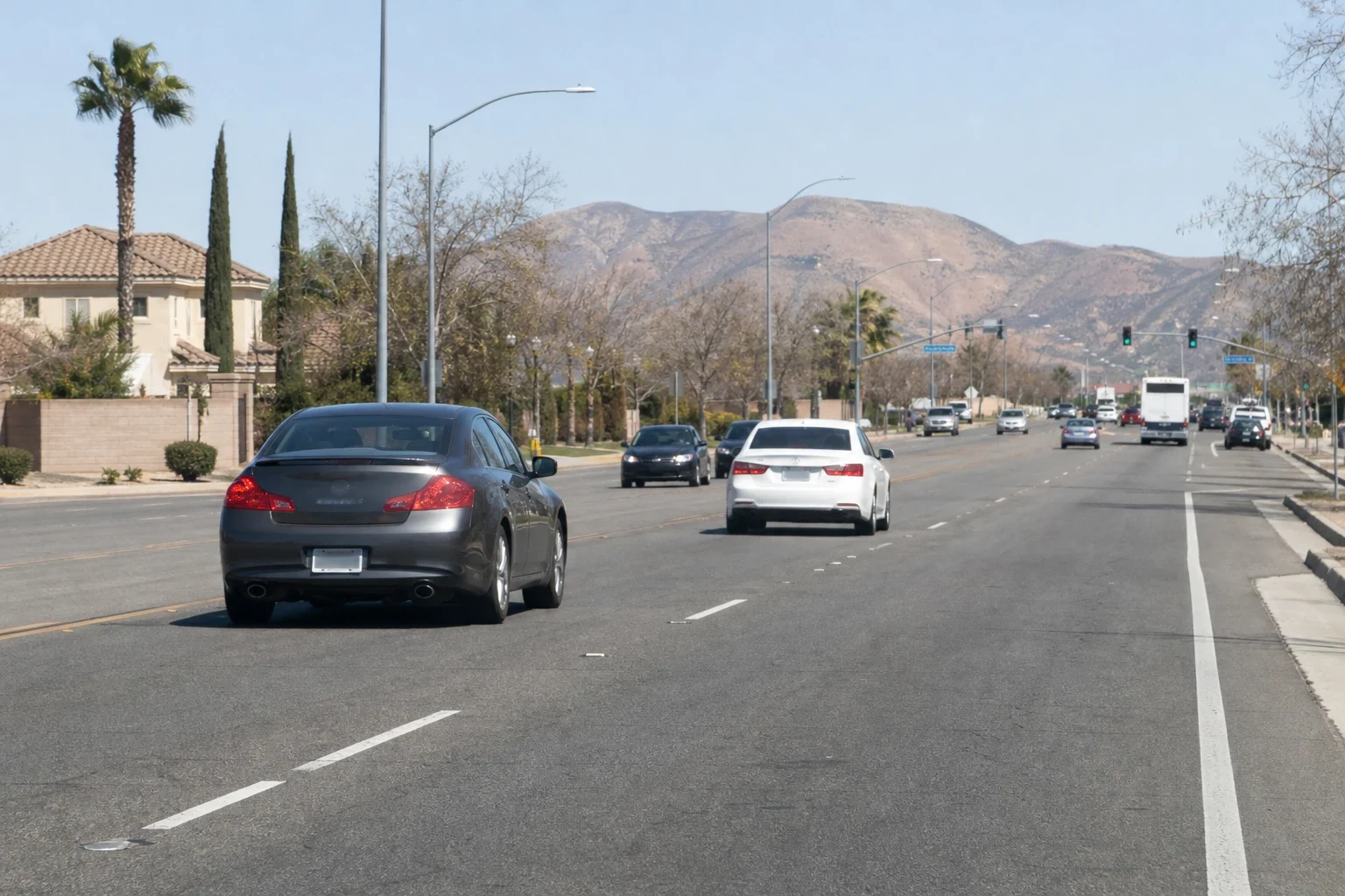 Wide suburban boulevard with cars driving past a stucco home, palm and cypress trees, and dry brown hills under a clear sky